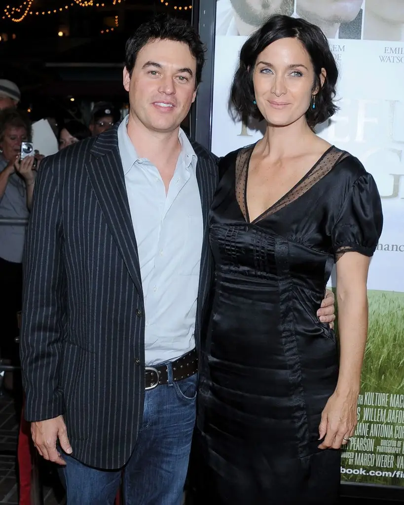 Carrie-Anne Moss, wearing a black dress, stands next to a man in a striped blazer and jeans.