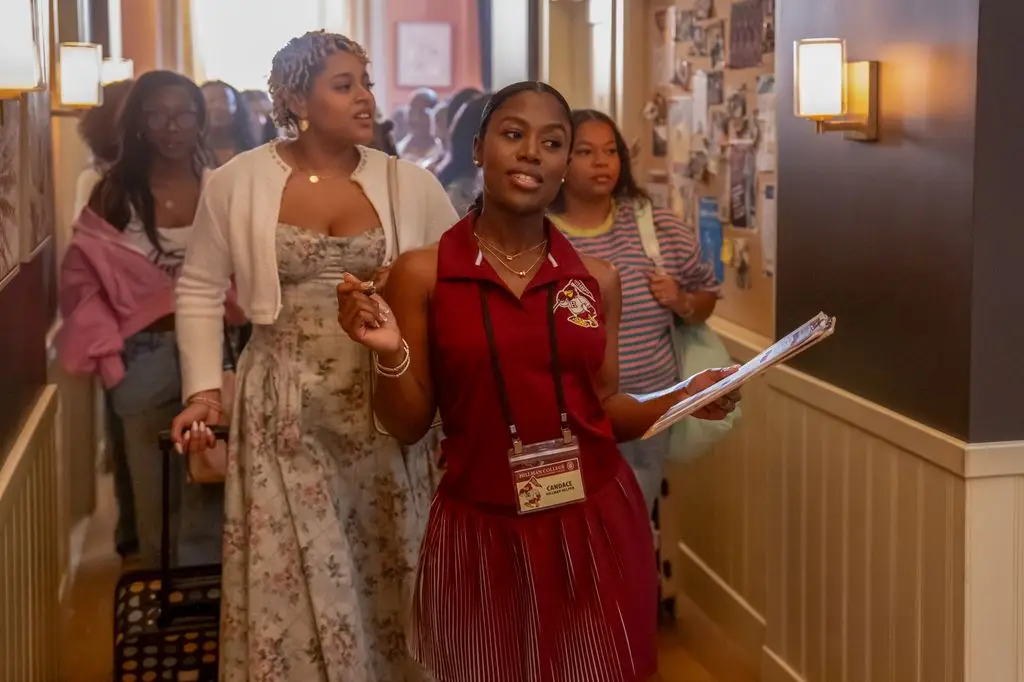 Group of women walking down a hallway, one in a red dress holding papers.