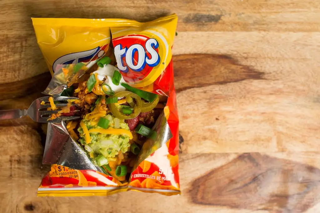 An opened bag of Fritos corn chips loaded with chili, cheese, sour cream, guacamole, jalapeños and spring onions, with a fork resting inside on a wooden board.