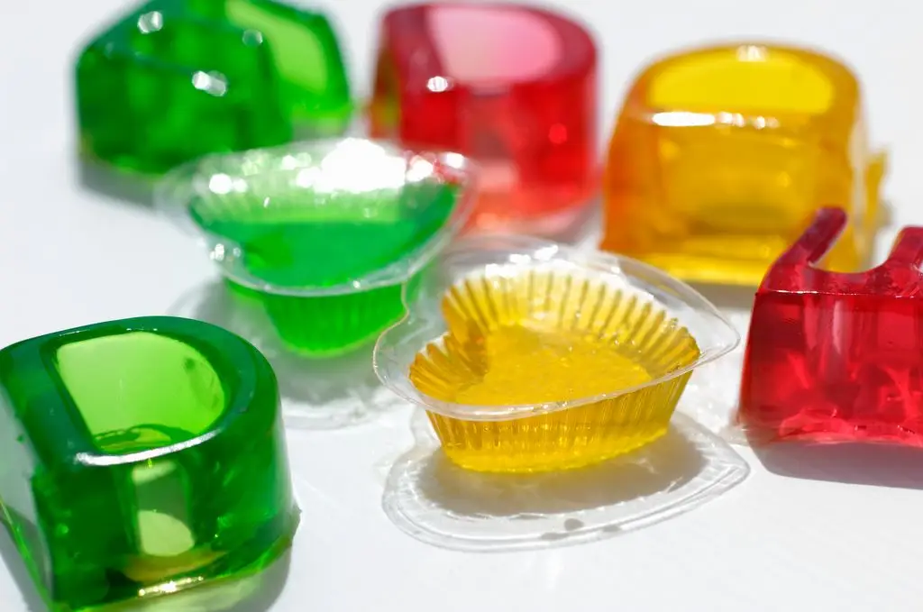 Several brightly colored jelly desserts in red, green and yellow, some still in their plastic molds, arranged on a white surface.