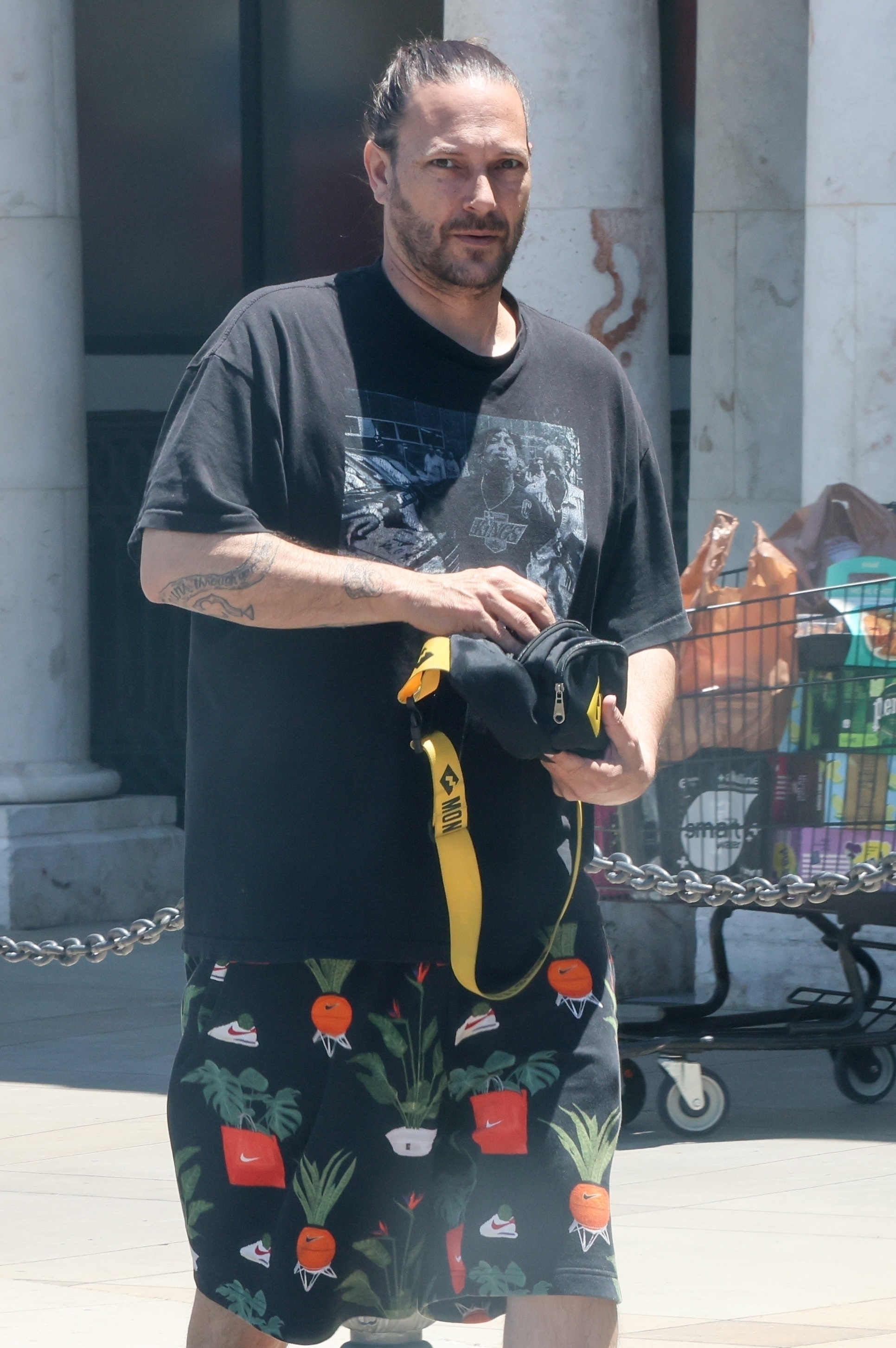 Kevin Federline wearing a black t-shirt and printed shorts while holding a black and yellow bag in front of a retail store.