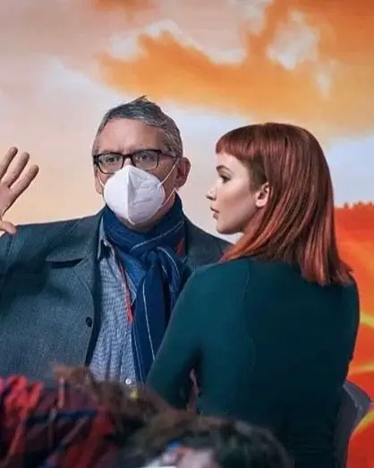 Adam McKay, wearing a mask and scarf, waves while talking to Jennifer Lawrence on a film set with an orange background.