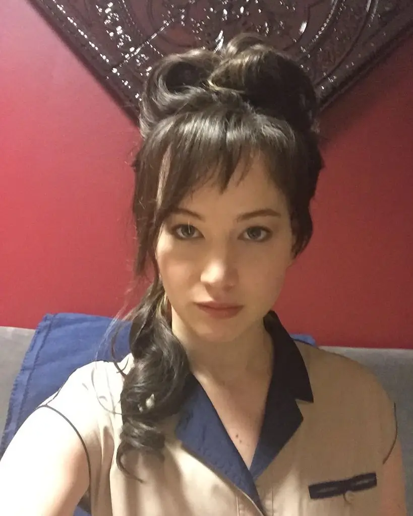 Jennifer Lawrence looks directly at the camera, her black hair styled in a voluminous bun with bangs, wearing a tan and navy collared top.
