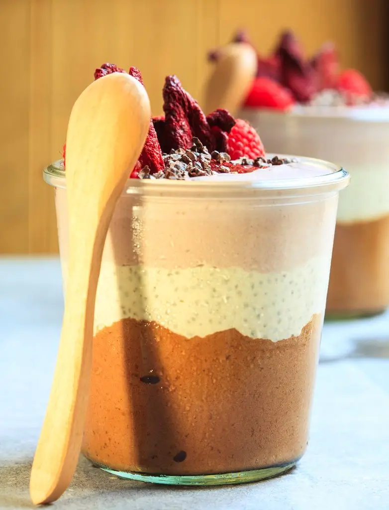 A layered dessert pot with layers of chocolate, cream and rose, garnished with raspberries, dried beetroot slices and cocoa nibs, with a wooden spoon placed on top.