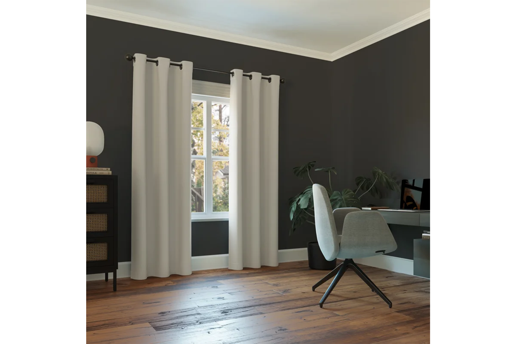 A room with dark walls and wooden floors, including a window with white curtains, a black cabinet, a potted plant, a desk and a gray office chair.