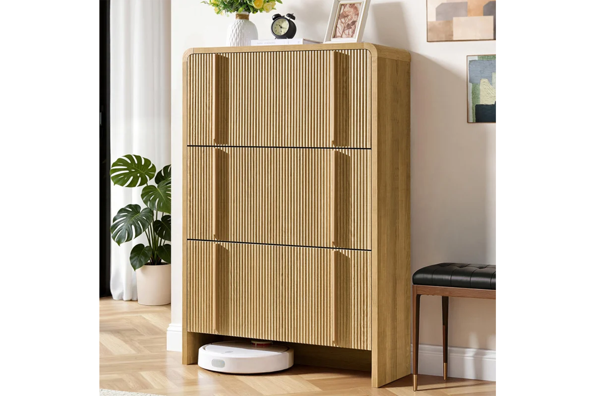 A light wood fluted shoe cabinet with three drop-leaf drawers stands against a white wall, topped with a vase of flowers and a clock, with a robot vacuum cleaner beneath.