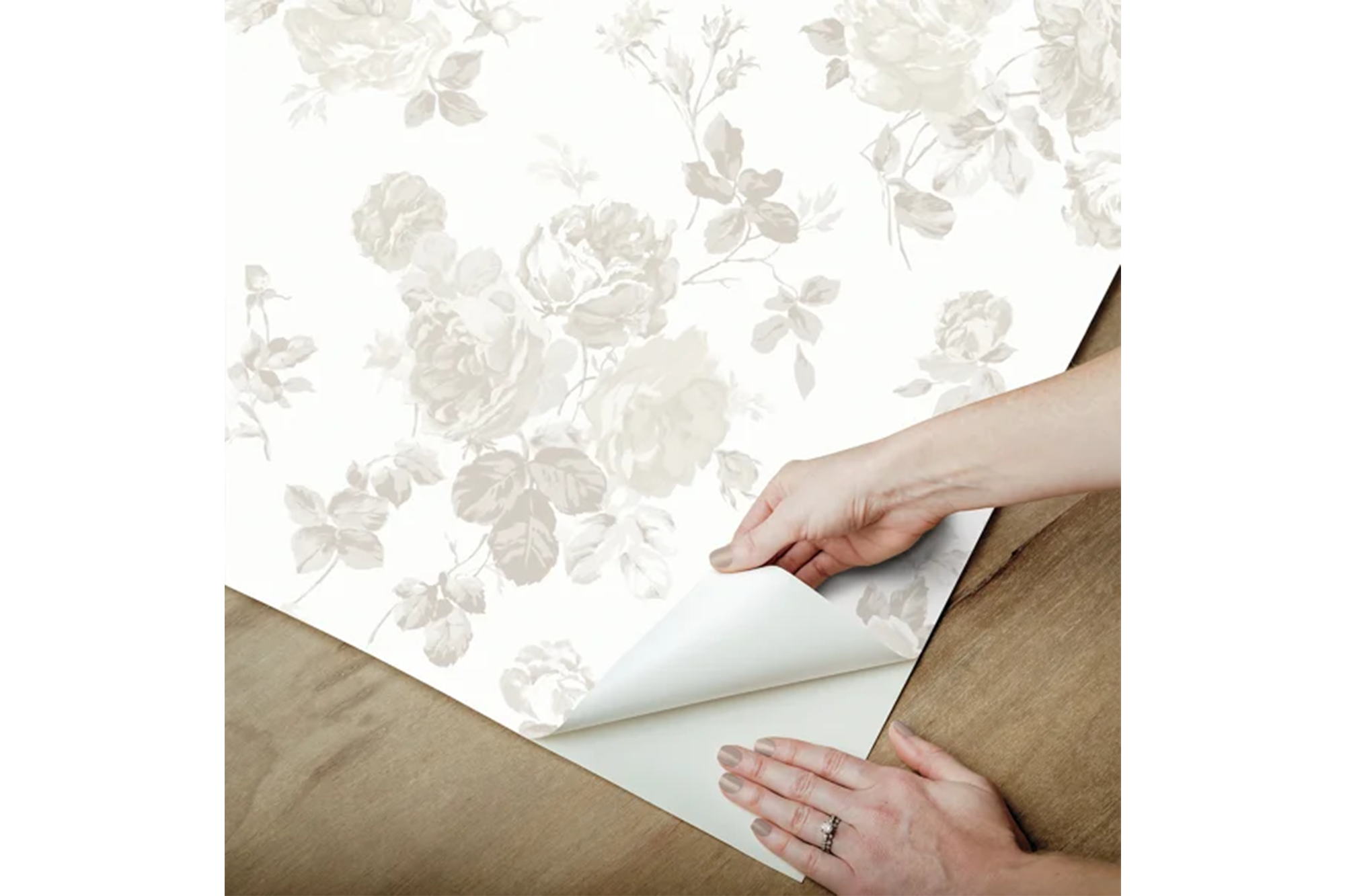 A pair of hands peel the backing off a sheet of white peel and stick wallpaper featuring a neutral floral pattern.
